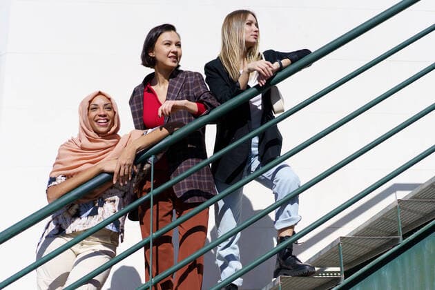 IMAGO Creative: Urban Diversity Content young diverse ladies looking away while standing on stairs in city Low angle of trendy young multiethnic female best friends, on stylish outfit and hijab, leaning on metal railing while standing on staircase and looking away in city on sunny day Copyright: xx 44186596