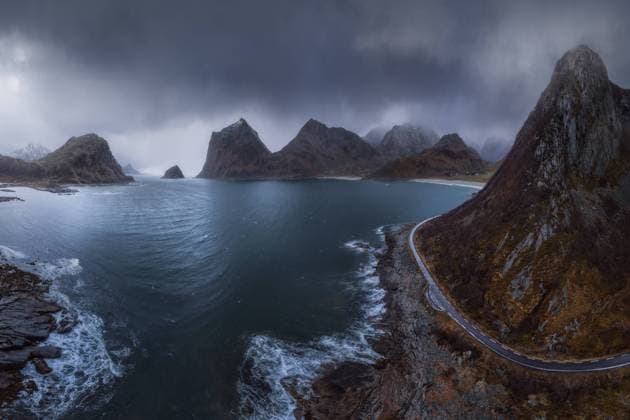 Sea with foamy waves surrounded by high rocky cliffs with road located against gray overcast sky in nature of Norway JuanLopez_LofotenDrone2_20.jpg Copyright: xJuanxLopezx Model Released