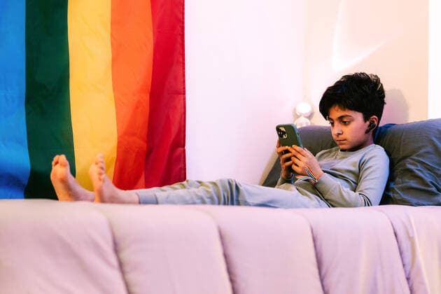 IMAGO Creative: Pride Month 2022, Engagement der Jugendlichen IMAGO Creative: Pride Month, Juni 2022 Full body side view of barefoot ethnic boy in wireless earbuds lying on bed and browsing cellphone near colorful LGBT flag HodeiUnzueta_YoungNonBinary_31.jpg Copyright: xHodeixUnzuetax Model Released