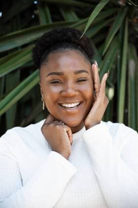 Creative Highlights Symbolbilder Positive African American female touching cheek while standing near lush green plants on summer day in garden DaniNavarro_BlackWoman_21.jpg Copyright: xDanixNavarrox Model Released