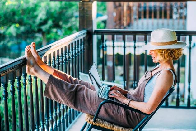 Portrait of a mature caucasian blonde young woman in her 50s with a laptop wearing a hat and sitting in a relaxed pose in a rocking chair on the wooden porch of a country house. Lifestyle concept. NAVARRE SPAIN Copyright: xMikelxBilbaox M-22-04-098