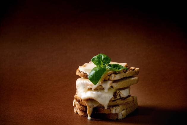 Stockpile of toasted melted cheese pressed sandwiches with basil leaves over dark brown background NatashaBreen REDA&CO 1028_29_NBR09955