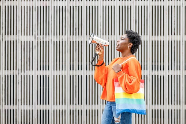 IMAGO Creative: Pride Month 2022, Aktivismus Side view of African American female with colorful bag talking in loudspeaker while giving speech on street near wall in city, Model released IgnacioFerrandiz_AfroStreet_62.jpg Copyright: xIgnacioxFerrandizx MR