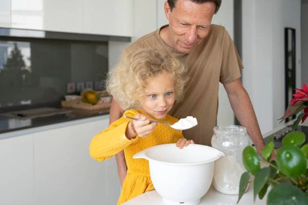 IMAGO Creative: Vaterschaft Smiling blond girl holding spoon of flour standing by father in kitchen model released, Symbolfoto property released, SVKF00140