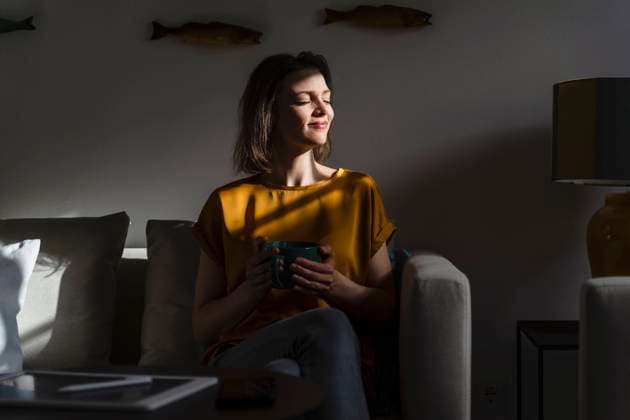 IMAGO Creative: Herbst, Gemütliches Zuhause Smiling woman with eyes closed holding coffee mug sitting in dark living room model released, Symbolfoto property released, DIGF17906