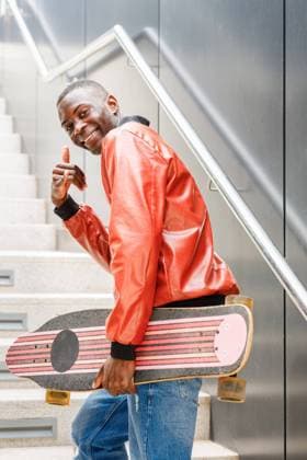 Side view of cheerful young African American guy in stylish outfit smiling at camera and showing call me gesture while walking upstairs with longboard in hand in city, Model released IgnacioFerrandiz_ZGenerationStudent_36.jpg Copyright: xIgnacioxFerrandizx MR