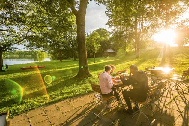 Menschen sitzen in der Abendsonne an einem Tisch einer Gartenwirtschaft und essen, Bayern, Deutschland. Gartenwirtschaft an einem Seeeinem kleinen See *** People sitting in the evening sun at a table of a garden restaurant and eating, Bavaria, Germany garden restaurant at a lakea small lake