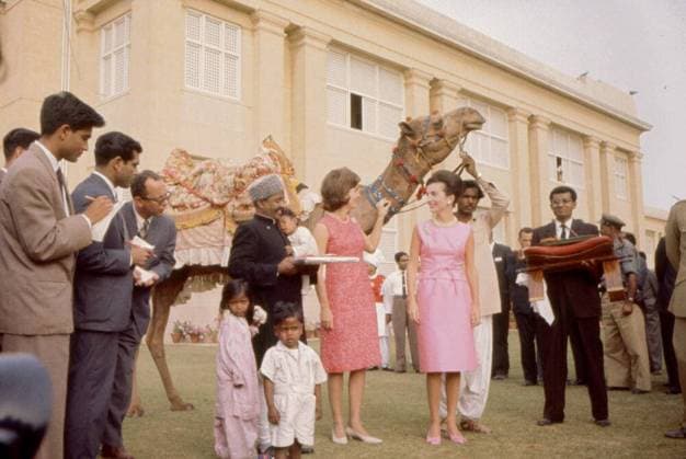 First Lady Jacqueline Kennedy s (JBK) trip to India and Pakistan: Karachi, Sindh, Pakistan, camel ride and visit with camel driver Bashir Ahmad and family. First Lady Jacqueline Kennedy (center) and her sister, Princess Lee Radziwill of Poland (right), visit with camel driver Bashir Ahmad (left of Mrs. Kennedy) and his family at the residence of President Mohammad Ayub Khan in Karachi, Sindh, Pakistan. Secret Service agent, Clint Hill, stands at right in background (partially hidden); all others are unidentified. Copyright: xpiemagsx