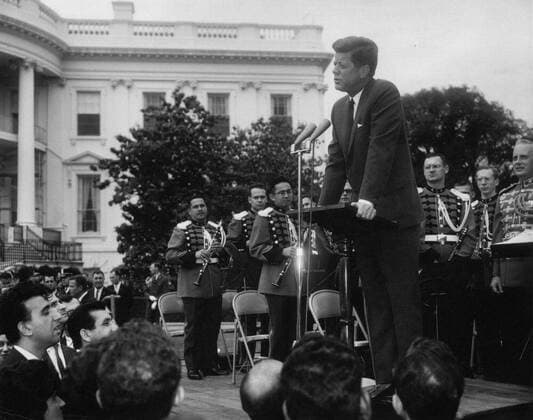Reception for Foreign Students, 3:55PM. President John F. Kennedy (at lectern) delivers remarks during a reception for foreign students on the South Lawn of the White House, Washington, D.C. Guests include students from the following Washington, D.C.-area institutions: American University, the Catholic University of America, Gallaudet College, the George Washington University, Georgetown University, Howard University, the Johns Hopkins University School of Advanced International Studies, Trinity College (Washington, D.C.), University of Maryland, and Dunbarton College of Holy Cross. Members of an unidentified band stand on platform; White House Secret Service agents, Ron Pontius and Emory Roberts, stand in background at left. Copyright: xpiemagsx