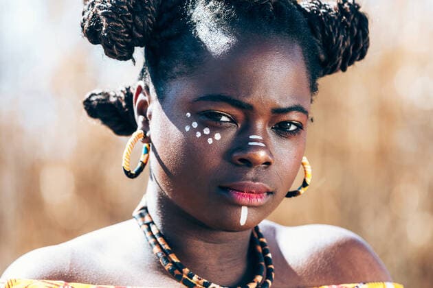 IMAGO Creative: UN Welt-Habitat-Tag am 3. Oktober Young black female in vivid authentic African dress with hair buns and traditional makeup folding hands on belly and looking at camera while standing on blurred background of sunlit field DavidBenito_AfricaPeople_27.jpg Copyright: xDavidxBenitox Model Released