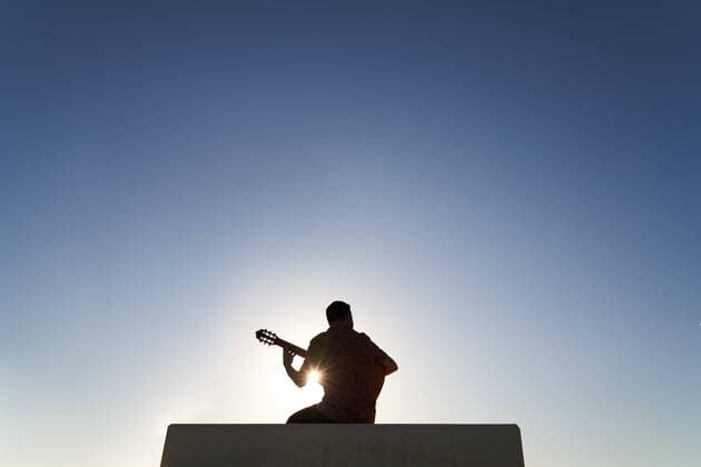 Back view silhouette of unrecognizable bearded musician playing song on acoustic guitar while sitting on border against blue cloudless sky on sunny day in city, Model released JavierBermudez_RubenGuitarrist_13.jpg Copyright: xJavierxBermudezx MR