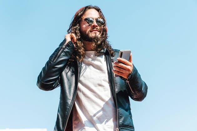 Bearded Latin American male in modern sunglasses and leather jacket browsing on cellphone and wireless headphones on blue sky background on sunny day, Model released MarcosCastillo_LatinCoupleLifestyle_7 Copyright: xMarcosxCastillox MR