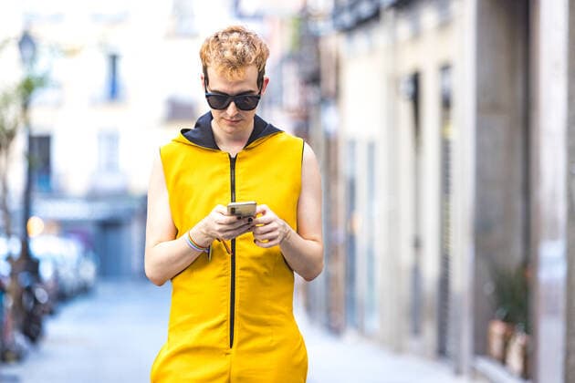 IMAGO Creative, Jahreszeit Sommer, Sommer in der Stadt Cheerful transgender young man in yellow jumpsuit and sunglasses looking at screen while walking and browsing on cellphone on city street DavidBenito_ID643_23435_024 Copyright: xDavidxBenitox Model Released