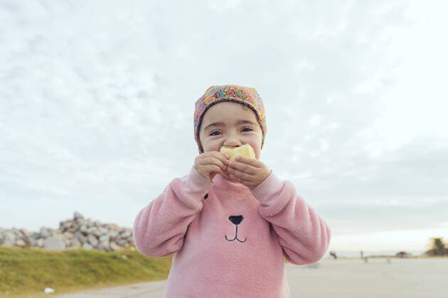 Creative Highlights Symbolbilder Cute Hispanic preschooler in pink sweatshirt and ornamental headscarf biting bun and looking at camera while standing on asphalt road against cloudy sky, Model released MathiasAlvez_ID503_19958_004 Copyright: xMathiasxAlvezx MR