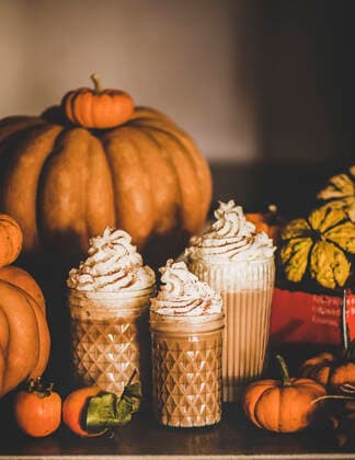 Pumpkin latte coffee with whipped cream in glasses on table Pumpkin spice latte coffee drink topped with whipped cream and cinnamon in tall glasses among fresh pumpkins and persimmons on table, grey wall at background. Seasonal Autumn hot warming sweet drink Copyright: xAnnaxIvanovax AI111119-3
