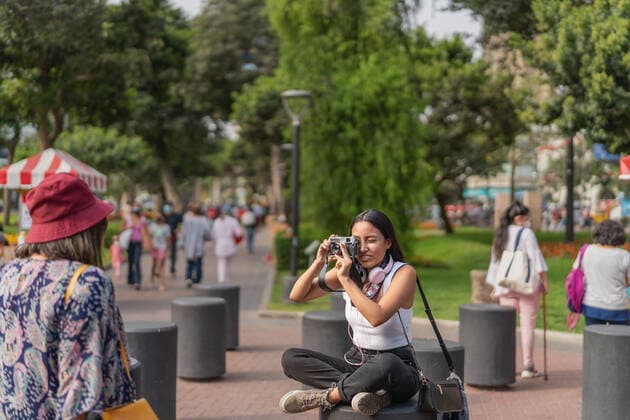 IMAGO Creative: Sommer, Sommertage im Stadtpark Peruvian female taking picture of anonymous female friend on photo camera while sitting on crowded street of city with trees, Model released GuillermoSpelucin_SaraiAndPaulaInLima_12 Copyright: xGuillermoxSpelucinx MR