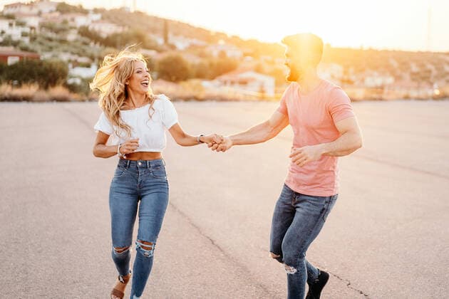 Cheerful man and excited woman looking at each other and laughing while walking dancing holding hands along asphalt road against blurred town on hillside in evening, Model released GustavoPozo_IsaAndKike_97 Copyright: xGustavoxPozox MR