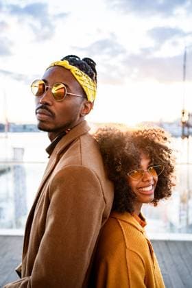 Creative Highlights Symbolbilder Side view of bright laughing black woman in yellow coat with trendy boyfriend in sunglasses standing back to back looking at camera on urban pier, Model released RafaCortes_AlphaDayanis_96 Copyright: xRafaxCortesx MR