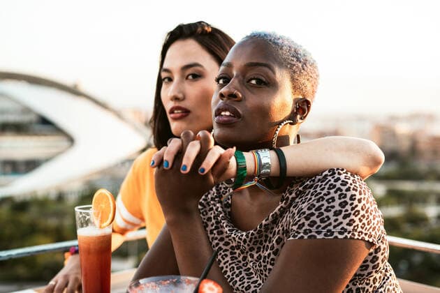 Stylish multiracial girlfriends sitting in open air cafe hugging and holding hands while enjoying refreshing drinks on blurred background, Model released RafaCortes_LesbianEthnicCouple_117 Copyright: xRafaxCortesx MR