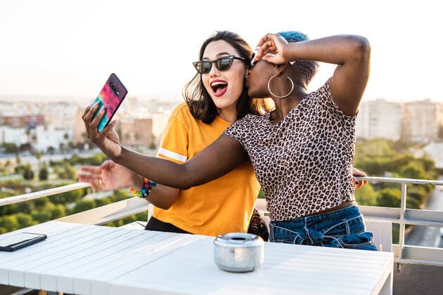Cheerful multiracial girlfriends in trendy sunglasses smiling sitting at table taking selfie on urban background, Model released RafaCortes_LesbianEthnicCouple_97 Copyright: xRafaxCortesx MR