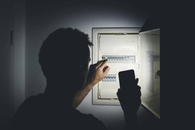 Bavaria, Germany - 25 September 2022: Power failure in the apartment, man shines the flashlight in his smartphone in the dark apartment and checks the fuse box. electricity, power outage, blackout concept *** Stromausfall in der Wohnung, Mann leuchtet mit der Taschenlampe in seinem Smartphone in der dunklen Wohnung und überprüft den Sicherungskasten. Elektrizität, Strom Ausfall, Blackout Konzept