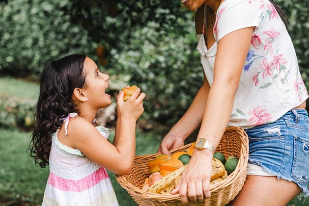 IMAGO Creative: Sommer, Sommertage im Garten Side view of cheerful Hispanic daughter smiling and eating carambola near crop mother with basket of fresh fruits on summer day in garden, Model released LeonardoBorges_MotherAndDaughterFieldDay_54 Copyright: xLeonardoxBorgesx MR