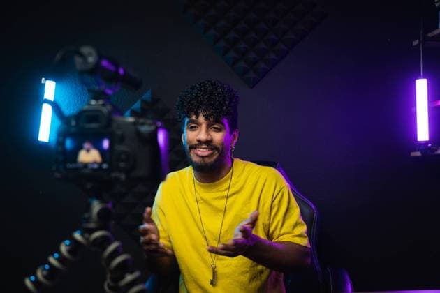 Optimistic young Hispanic male vlogger in yellow t shirt smiling and talking to audience while sitting at table in front of camera and recording video for blog in dark studio PabloMenendez_ID10507_318325_011 Copyright: xPabloxMenendezx Model Released