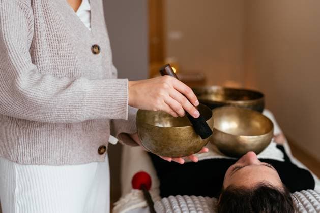 Side view of anonymous female sound massage therapist in uniform standing near client with Tibetan singing bowl while doing procedure in spa salon AidaMartinez_ID12552_381045_008 Copyright: xAidaxMartinezx Model Released