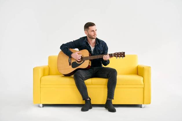 Handsome young man playing acoustic guitar whlile sitting on yellow sofa over white background Handsome young man playing acoustic guitar whlile sitting on yellow sofa over white background. Hobby, music concept Copyright: xZoonar.com ViktorxGladkovx 18794796 ,model released, Symbolfoto ,property released