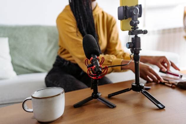 Anonymous black female blogger in casual clothes sitting on sofa and organizing makeup supplies near cup of coffee and microphone while preparing for recording podcast on smartphone placed on tripod JakeJakab_ID13042_396953_003 Copyright: xJakexJakabx Model Released