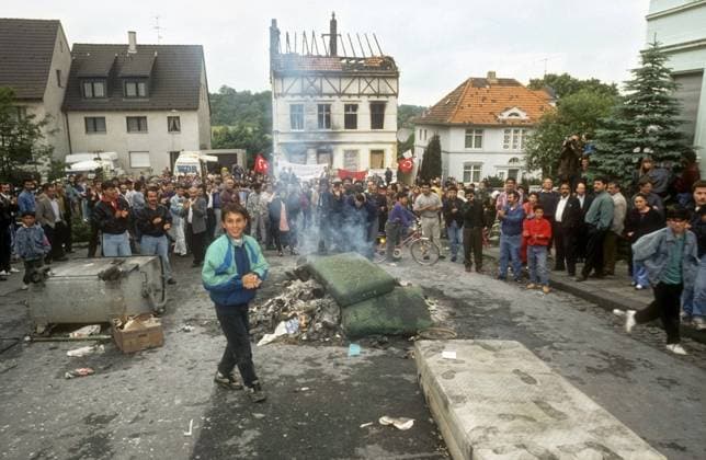 Solingen, Deutschland, DEU - Rassistischer Brandanschlag auf das Haus der tuerkischstaemmigen Familie Genc mit 5 Opfern (29.5.1993) 31.05.1993, Solingen, Deutschland, DEU - Rassistischer Brandanschlag auf das Haus der tuerkischstaemmigen Familie Genc mit 5 Opfern (29.5.1993). Zwei Tage nach dem rassistischen Brandanschlag auf das Haus der tuerkischstaemmigen Familie Genc am 29.5.1993. Fuenf Frauen und Maedchen verbrannten und erstickten. Tuerkischstaemmige Maenner protestieren beim Tatort. Im Hintergrund die Ruine des Brandanschlages. Der Anschlag war der traurige Hoehepunkt einer deutschlandweiten Welle rassistischer Angriffe auf Fluechtlinge und Migranten. Die Taeter waren vier junge Maenner der oertlichen Neonaziszene zwischen 16 und 23