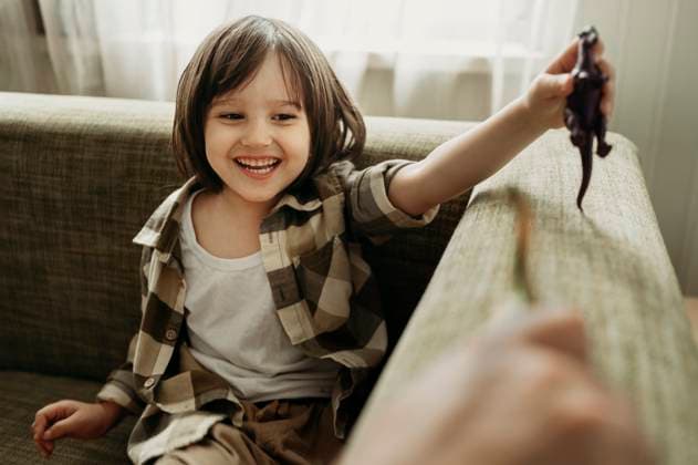Cheerful son playing with toy dinosaur on sofa at home model released, Symbolfoto property released, ANAF01408