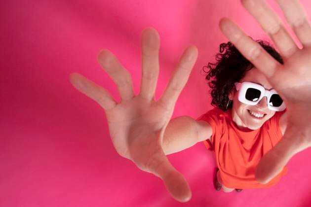 MR_ID13554_13235_AgustinaGates.jpg From above of cheerful young female in bright outfit and white sunglasses raising arms with palms at camera on pink background, Model released DondykRiga_ID13554_412012_030 Copyright: xDondykxRigax MR