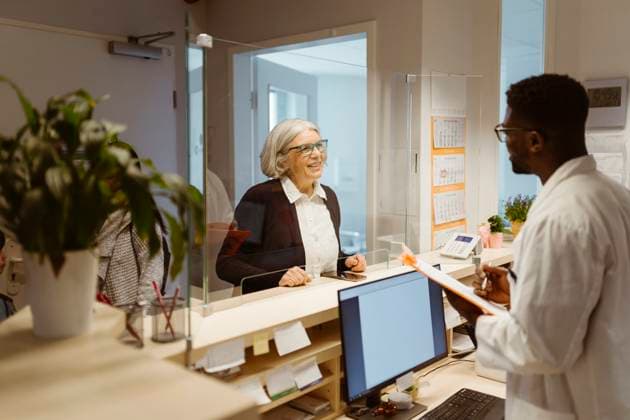Smiling senior female patient communicating with male receptionist through transparent shield in clinic Germany, Berlin, model released, property released Copyright: xMaskotx , MA101288 MA101288