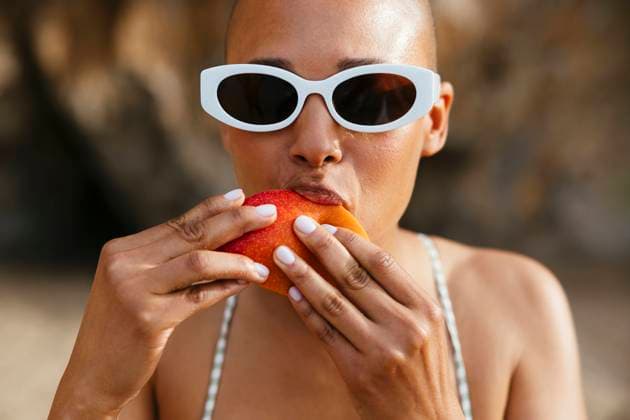 Creative Highlights Symbolbilder Woman wearing sunglasses eating mango at beach model released, Symbolfoto, EBSF03364