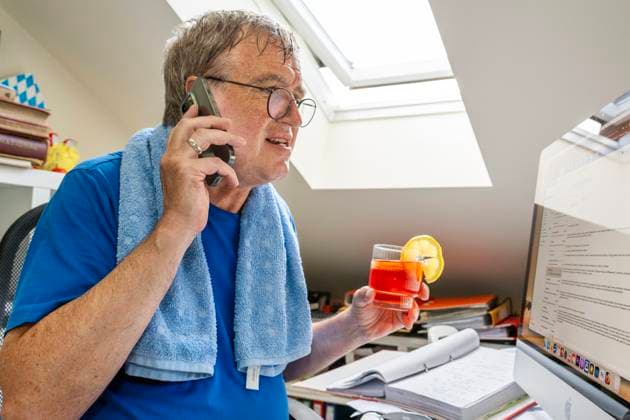 IMAGO Creative, Jahreszeit Sommer, Hitze Mann schwitzt im Home Office, leidet unter Rekordhitze in der Dachwohnung, München, Juli 2023 Deutschland, München, Juli 2023, Mann schwitzt im Home Office, leidet unter Hitze in der Dachwohnung, gönnt sich ein kühles Getränk, Glas Aperol, sitzt unter der Dachschräge, Sonne knallt durch das Fenster ins Büro, Schweiß läuft ihm übers Gesicht, er telefoniert und arbeitet am Computer, Alkohol in der Arbeitszeit, Handtuch liegt um die Schultern, schwitzen, Dachfenster geöffnet, Brille, Wohnung, Bayern, *** Man sweating in home office, suffering record heat in rooftop apartment, Munich, July 2023 Germany, Munich, July 2023, Man sweating in home office, suffering record heat in rooftop apartment, allows himself a cool drink, glass of Ap