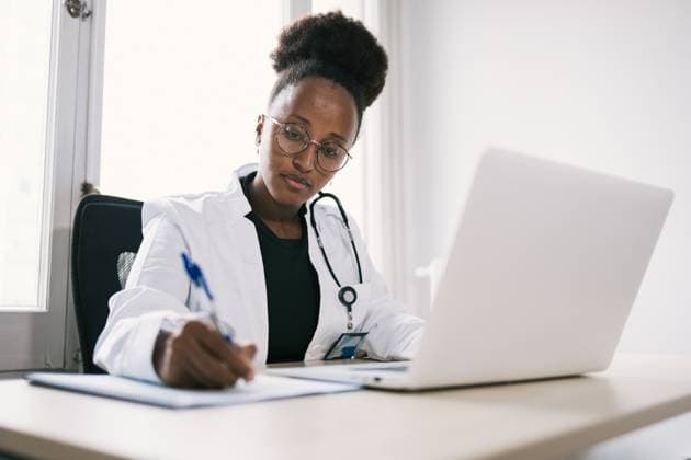MR_ID14857_13901_TsionRodriguez.jpg, PR_ID14857_3900_Pr-aetna.jpg Concentrated African American female in medical uniform sitting at table with laptop and writing on paper, Model released, Property released DavidMunoz_ID14857_453373_005 Copyright: xDavidxMunozx MR