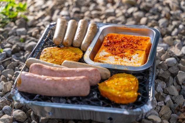 Bavaria, Germany - 2 September 2023: Vegetarian Grilling, Bratwurst Vegan and Vegetarian on a disposable grill on pebbles next to a meadow. Meat and sausage substitutes *** Vegetarisch Grillen, Bratwürste Vegan und Vegetarisch auf einem Einweggrill auf Kieselsteinen neben einer Wiese. Fleisch und Wurst Ersatzprodukte