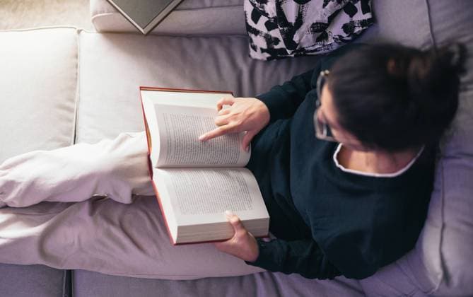 Young woman sitting on sofa and reading book in daylight From above of young female with eyeglasses looking down while relaxing on sofa cum bed with index finger on paper of reading book in daylight at home Model Released EricPanades_ID16559_503251_029 Copyright: xEricxPanadesx RECORD DATE NOT STATED