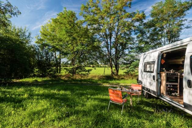 Ein Camper steht zwischen Bäumen an einem sonnigen Sommertag auf einer Wiese im Grünen, ein gedeckter Frühstückstisch mit Stühlen steht vor der Schiebetür des Campers. Regensburg, Bayern, Deutschland. In der Altstadt von Regensburg *** A camper stands between trees on a sunny summers day on a meadow in the countryside, a laid breakfast table with chairs stands in front of the sliding door of the camper Regensburg, Bavaria, Germany In the old town of Regensburg