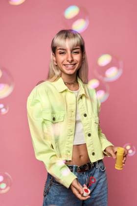 Creative Highlights Symbolbilder Young cheerful blond haired female with face piercings looking at camera in yellow jacket blowing soap bubbles from bottle while standing against pink background Model Released ManuelRuiz_ID16291_495899_018 Copyright: xManuelxRuizx
