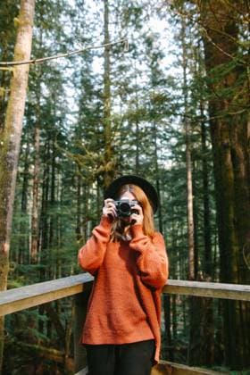Creative Highlights Symbolbilder A woman in a warm orange sweater and hat stands on a bridge in the lush green forests of Avatar Grove, Vancouver Island, holding a camera to her eyes. Model Released CamilleGirard_ID17696_535283_031 Copyright: xCamillexGirardx
