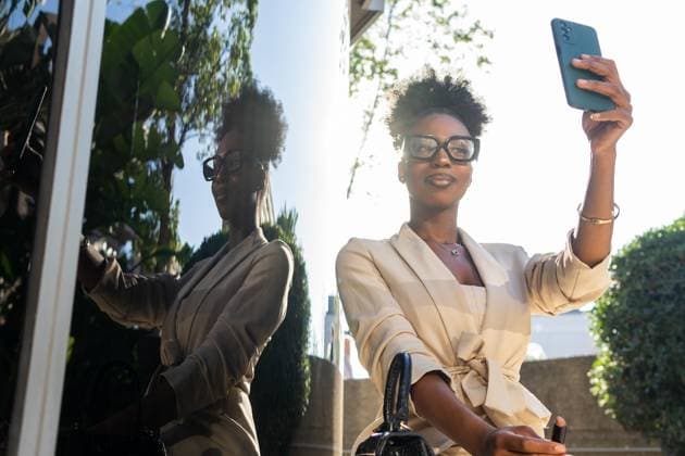 Low angle of beautiful young black female manager in formals and eyeglasses with lipstick sitting by glass wall and looking at smartphone while taking selfie Model Released DavidMunoz_ID17348_527732_020 Copyright: xDavidxMunozx