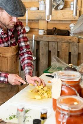 IMAGO Creative, Food, Trend, Meal Prep Crop focused chef in apron and beret standing in a rustic kitchen attentively dicing potatoes on a white cutting board with a background of hanging utensils and jars of homemade preserves Model Released JuanAizpuru_ID18273_546351_022 Copyright: xJuanxAizpurux