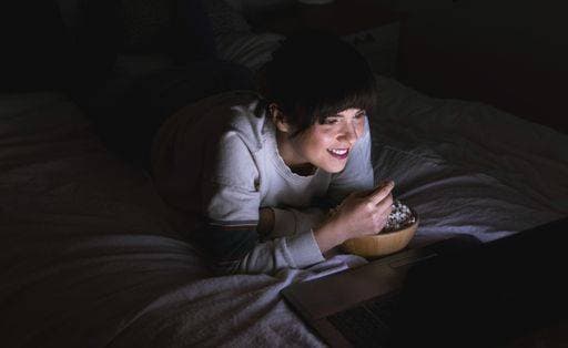 IMAGO Creative, Weihnachten, Tradition: Weihnachtsfilme sehen A female lying on her bed, watching a movie on her laptop and eating popcorn in the darkness 1168088 ,model released, Symbolfoto RECORD DATE NOT STATED