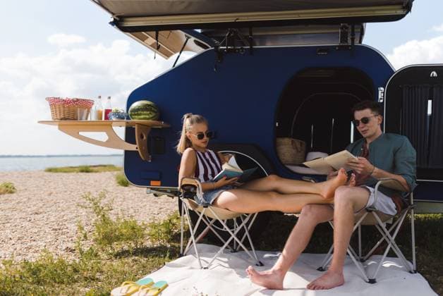 Young couple sitting together in front of van, camping and reading book. Happy young couple sitting together in front of van, camping and reading a book. model released Copyright: xx SUMMER WATER TRIP COUPLE 20222