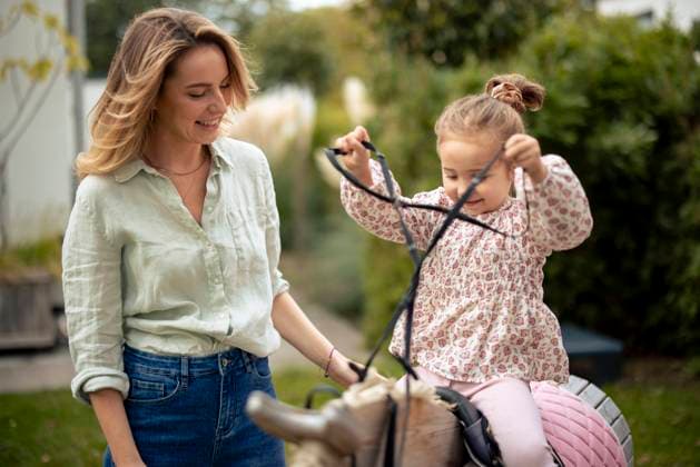 IMAGO Creative, Kinder auf dem Reiterhof Happy girl playing on wooden horse near mother in garden model released, Symbolfoto property released, JOSEF23173