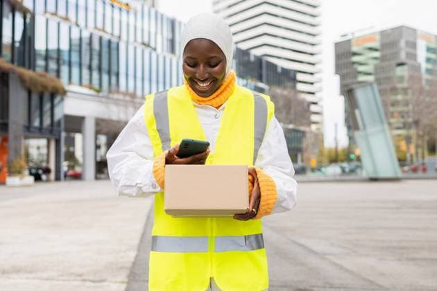 Smiling Muslim African woman in hi-vis jacket using her smartphone while holding a parcel on a city street, radiating positivity and professionalism. Model Released DavidMunoz_ID20424_604009_005 Copyright: xDavidxMunozx