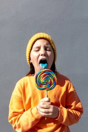 Creative Highlights Symbolbilder A young individual in an orange sweatshirt and yellow beanie savors the taste of a large, vibrant swirl lollipop against a plain background Model Released SergioVictorVega_ID20390_603179_009 Copyright: xSergioxVictorxVegax