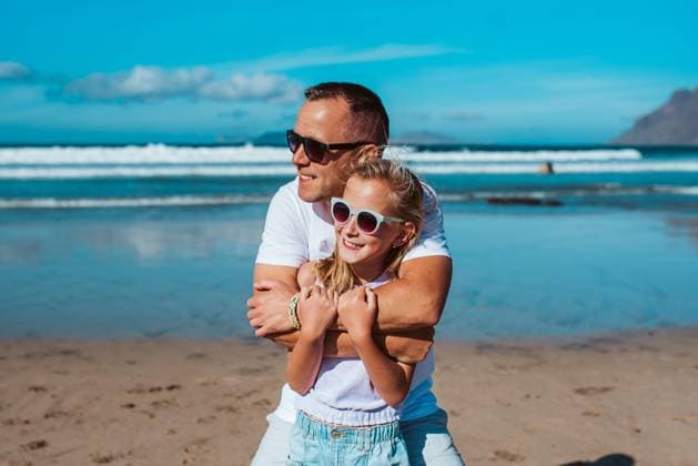 Father embracing daughter on beach. Dad and girl enjoying sandy beach, looking at crystalline sea in Canary Islands. Concept of beach summer vacation with kids. Father embracing daughter, standing on beach. Dad and girl enjoying sandy beach, looking at crystalline sea in Canary Islands. Concept of beach summer vacation with kids. model released